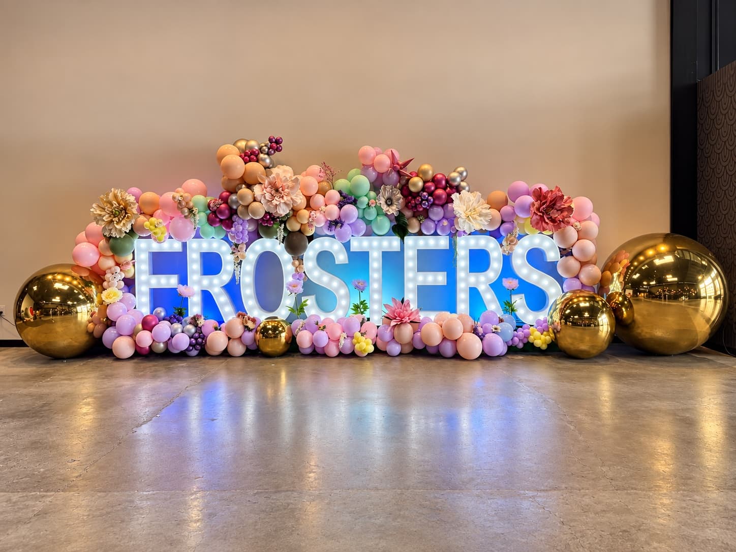 An illuminated marquee-letter corporate event installation framed by a pastel balloon and floral garland with gold mirror spheres, designed by Events by Juna.