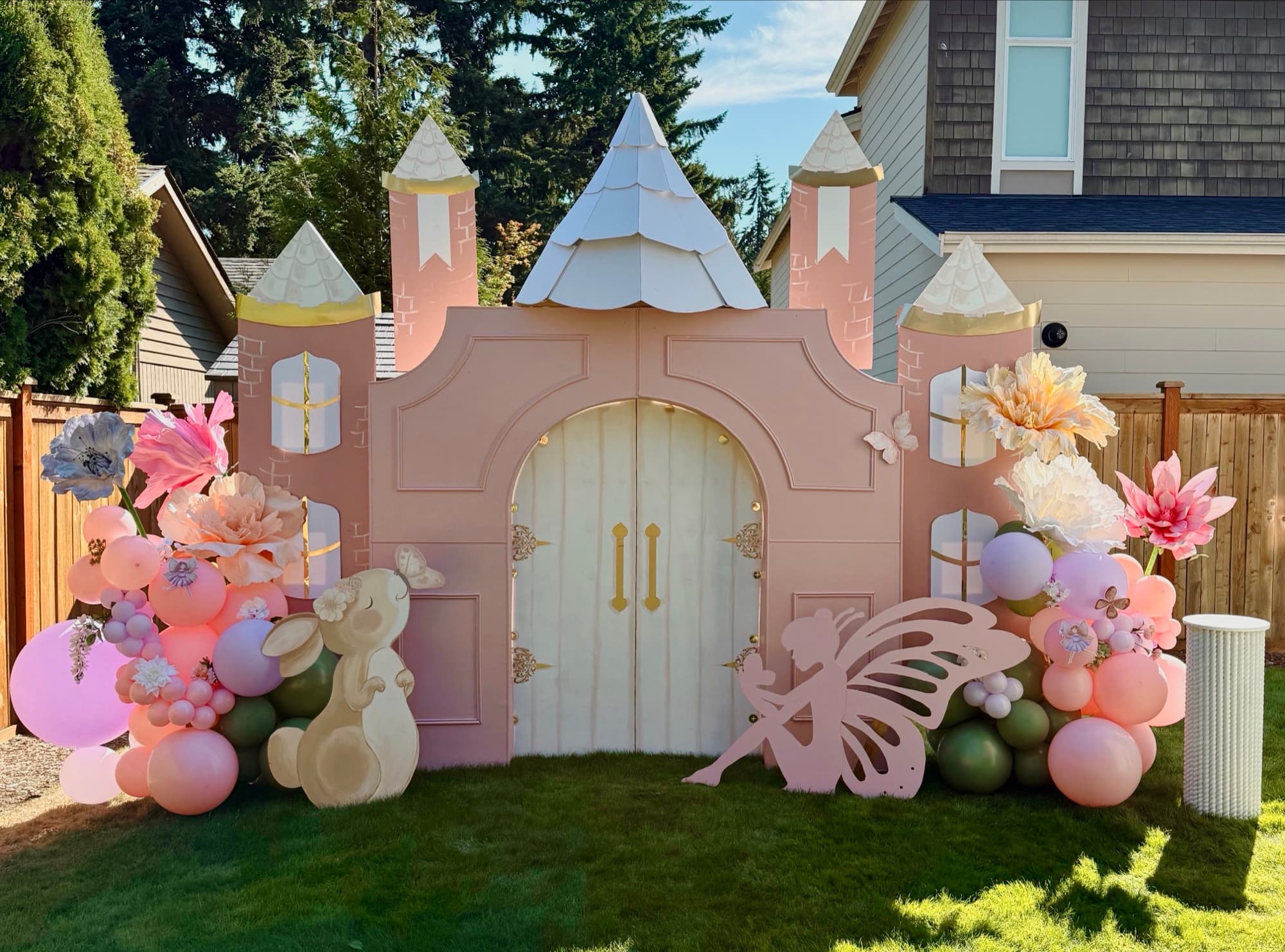 A pastel pink fairy-tale castle backdrop with oversized paper florals and a balloon garland, designed by Events by Juna for a children's celebration.