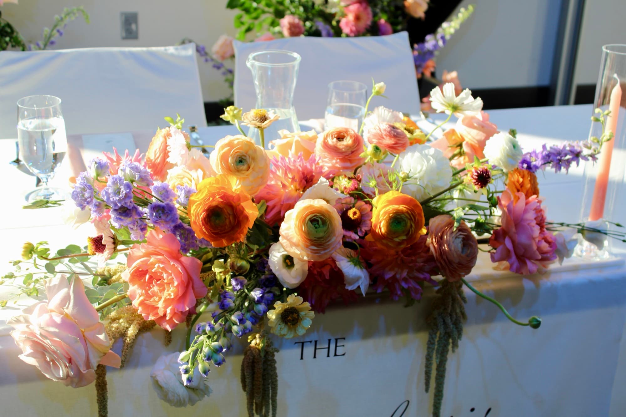 A lush sweetheart-table floral garland in peach, coral and lavender with trailing greenery, designed by Events by Juna for a wedding.
