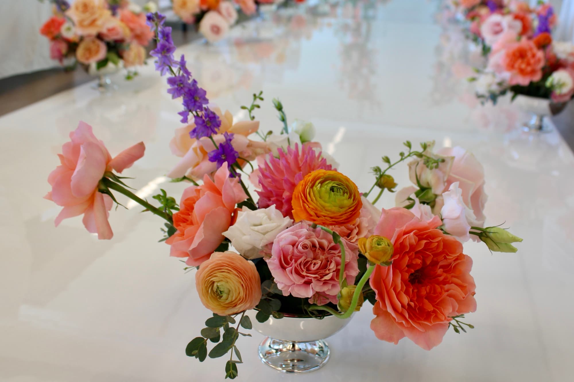 A footed silver-bowl wedding centerpiece of peach garden roses, ranunculus and purple delphinium by Events by Juna.